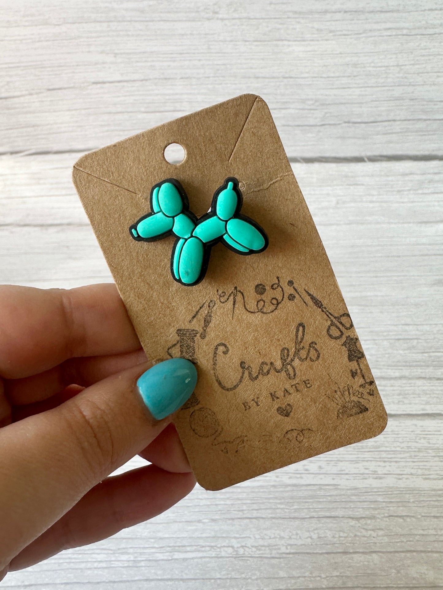 A hand with turquoise nails holds a brown kraft card labeled "Crafts by Kate," featuring crafting tool illustrations and displaying the Ball is Life - Arnie & Mac’s Goodies pin—a blue balloon dog, perfect for decorating your pet's bandana. White wooden background.