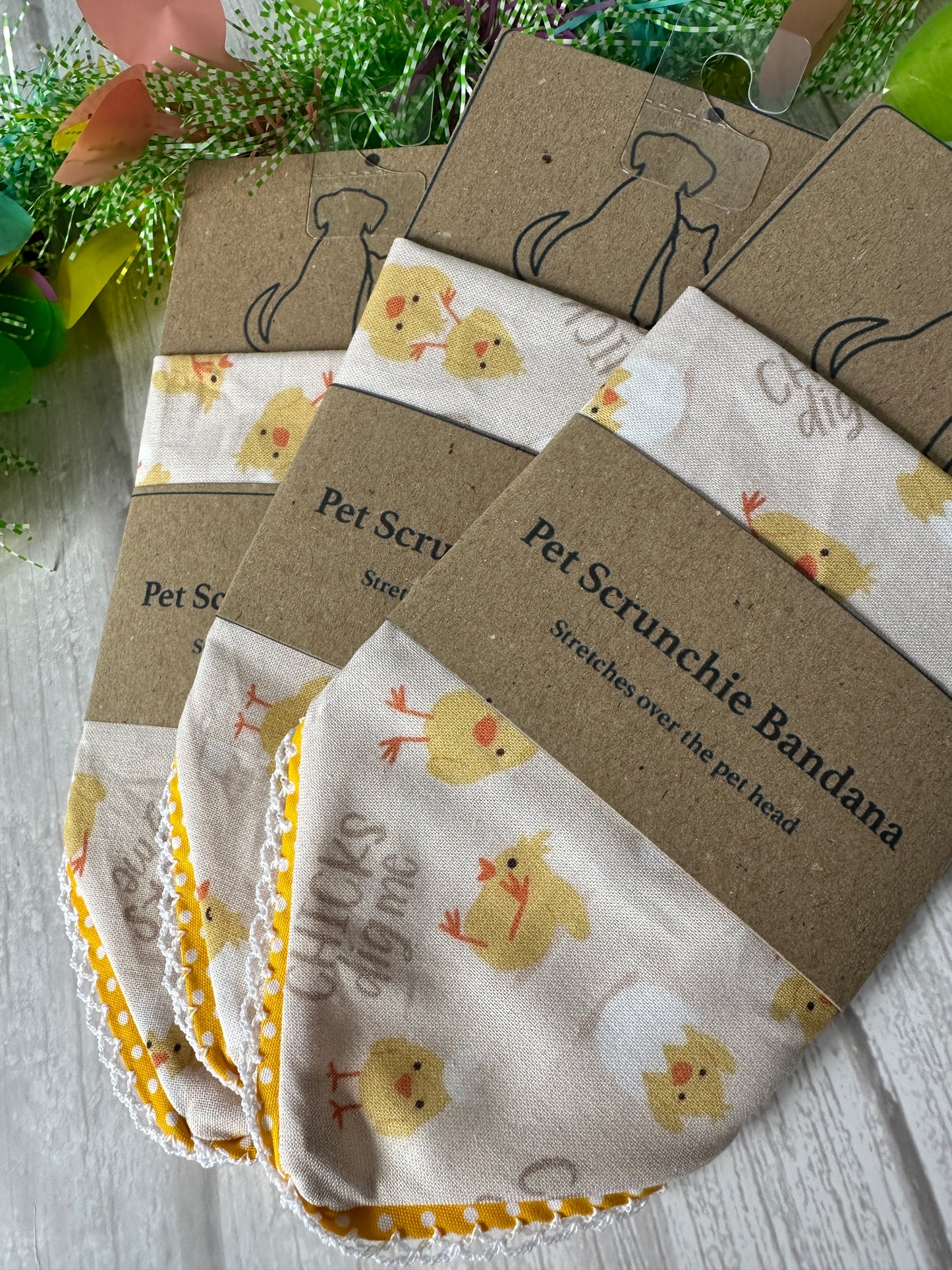 Crafts by Kate's Cotton Pet Scrunchie Bandana - Chicks Dig Me features cute chick designs on a tabletop. The packaging displays a cat outline and the phrase "Pet Scrunchie Bandana" set against a floral, spring-themed backdrop, ideal for Barkday celebrations.