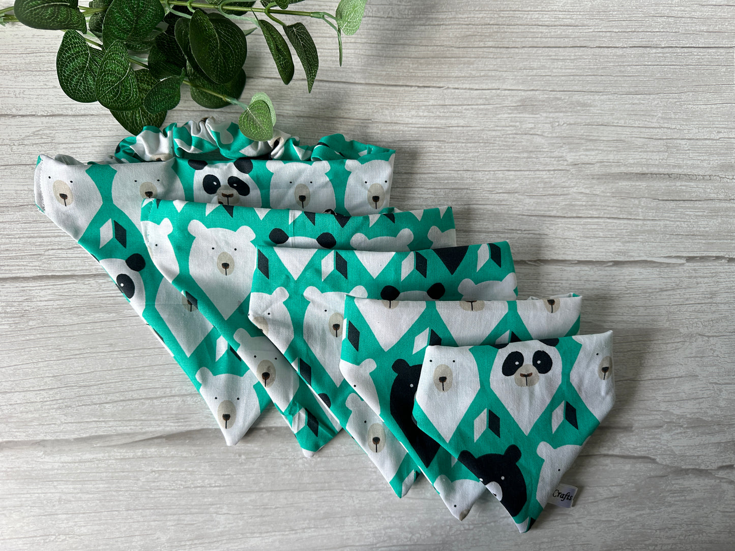 A set of four triangular cotton "It’s My Barkday" bandanas by Crafts by Kate are arranged in a fan shape on a light wooden surface. The green bandanas, adorned with cartoon pandas, are perfect for Bear Lovers Fabric enthusiasts, with green leaves visible at the top left.