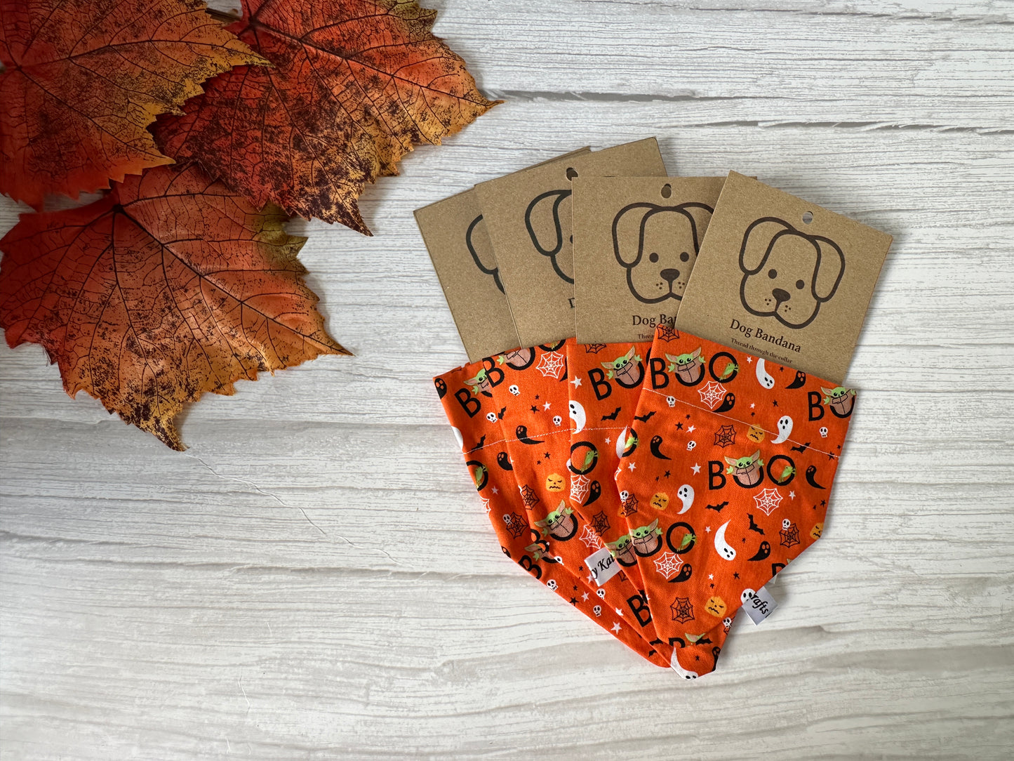 Three orange Cotton Pet Bandanas with collar attachment by Crafts by Kate, featuring Halloween Baby Yoda designs, are displayed on a light wooden surface with autumn leaves. Each has a brown tag with a cartoon dog face.