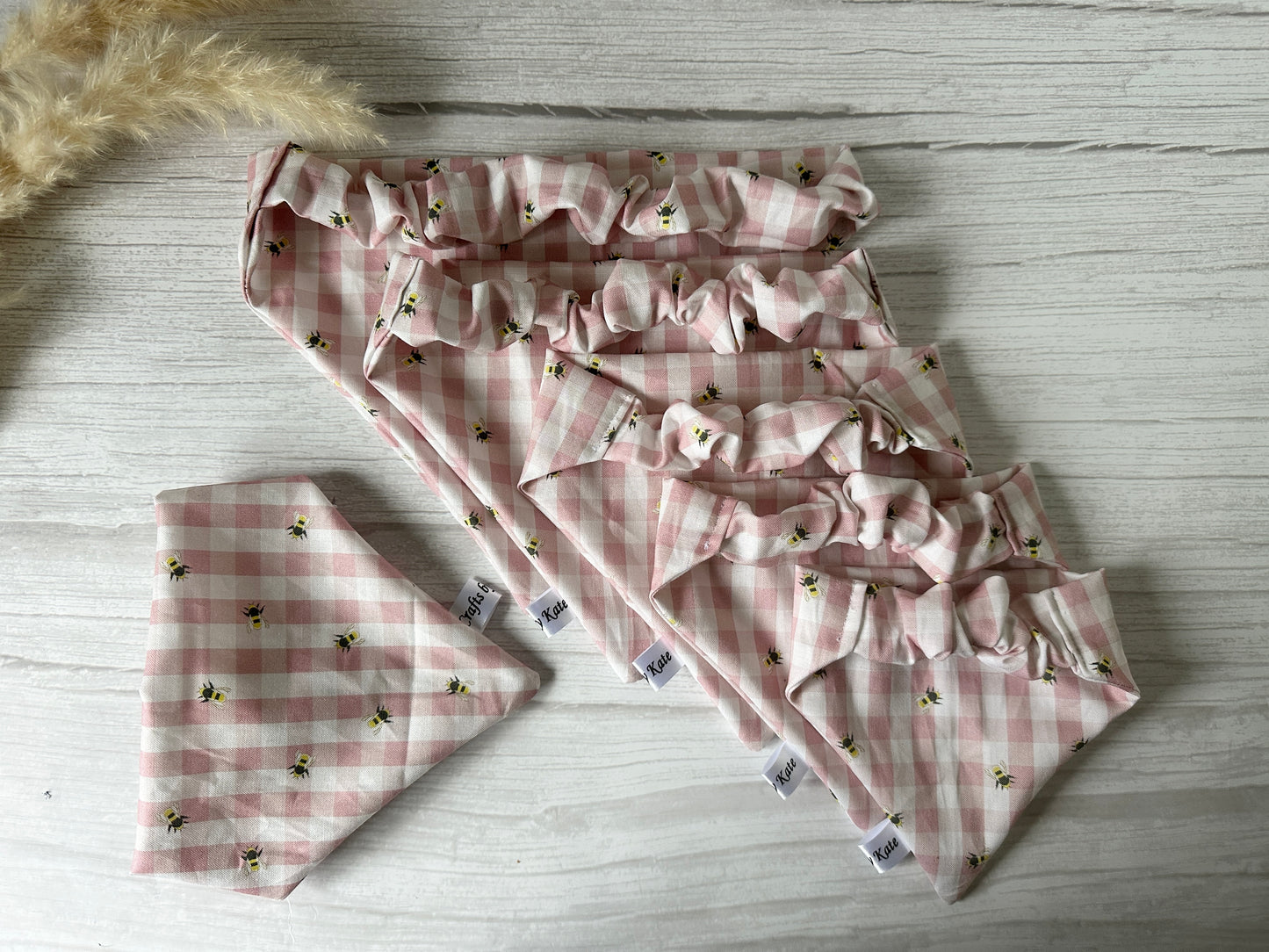 Five Crafts by Kate pink gingham bees fabric dog bandanas are on a white wooden surface with a matching Cotton Pet Scrunchie Bandana on the left. Beige pampas grass decorates the top left corner, adding texture to the cozy scene.