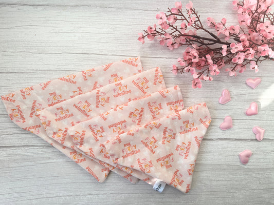 Crafts by Kate's handcrafted "My Fluffy Valentine" cotton pet bandanas with collar attachments are beautifully displayed on a light wooden surface, adorned with fluffy heart-shaped decorations and soft pink blossom branches.