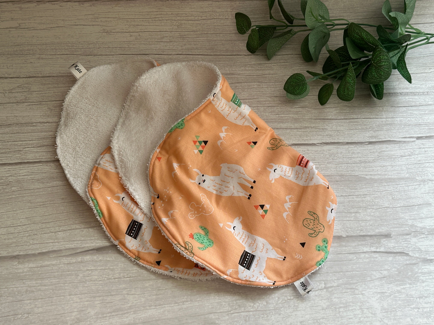 Two "Llama Loves" cotton baby bibs by Crafts by Kate, featuring llama and cactus patterns on a peach background, rest on a light wooden surface. A small green plant in the top right corner adds a natural touch to this soothing scene.
