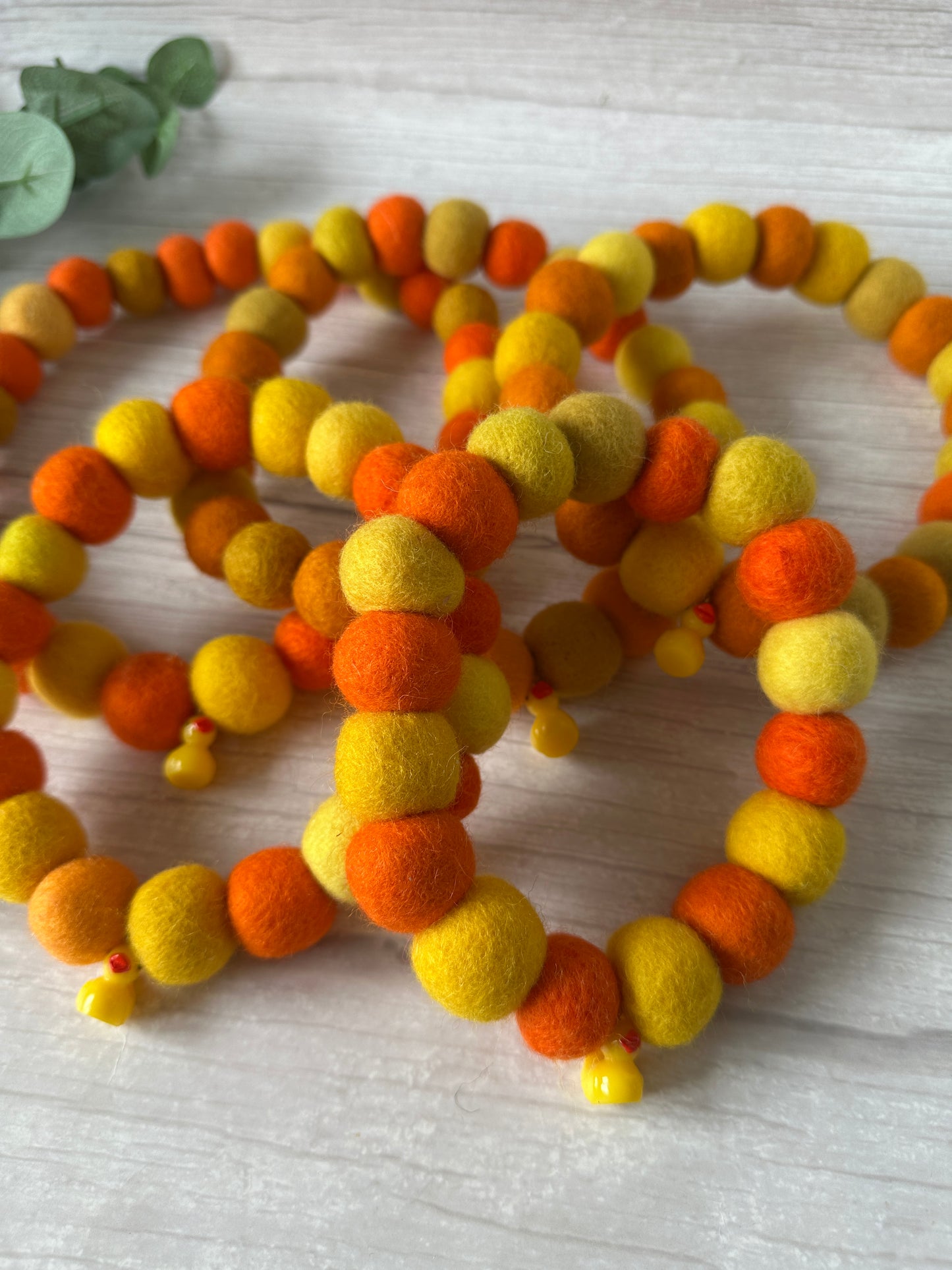 Three PomPom Pet Collar - Rubber Ducks by Crafts by Kate, handcrafted from round felt balls in yellow, orange, and red, are displayed on light wood. Each collar has a small yellow bead at one end. Green leaves appear in the corner.