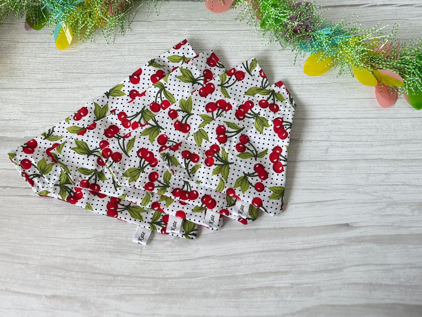 The Cotton Pet Bandana with collar attachment from Crafts by Kate's Cherry Sweet collection features a cherry pattern on white polka dots, atop a light wooden surface, complete with colorful tinsel in the upper corner.