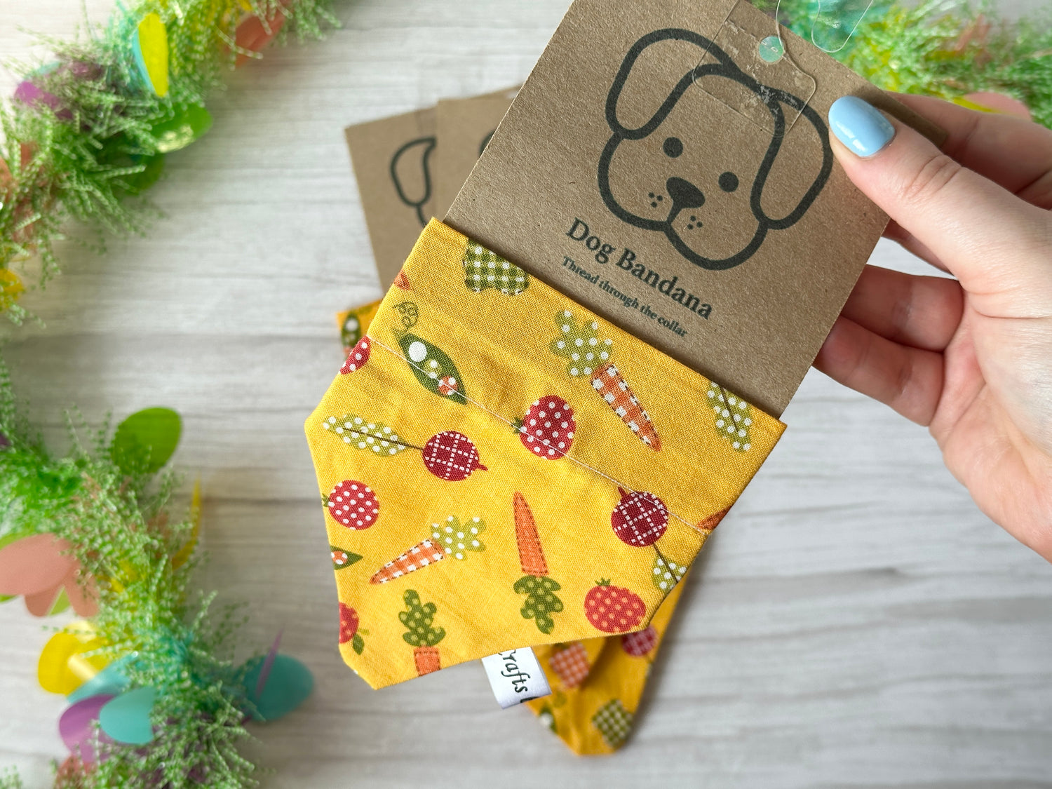 A hand holds a packaged, handcrafted "Cotton Pet Bandana with collar attachment - Vegetable Patch" by Crafts by Kate. It features vibrant vegetable patterns like carrots and broccoli on yellow fabric, adorned with a cartoon dog face and floral border, sliding effortlessly through the pet's collar.