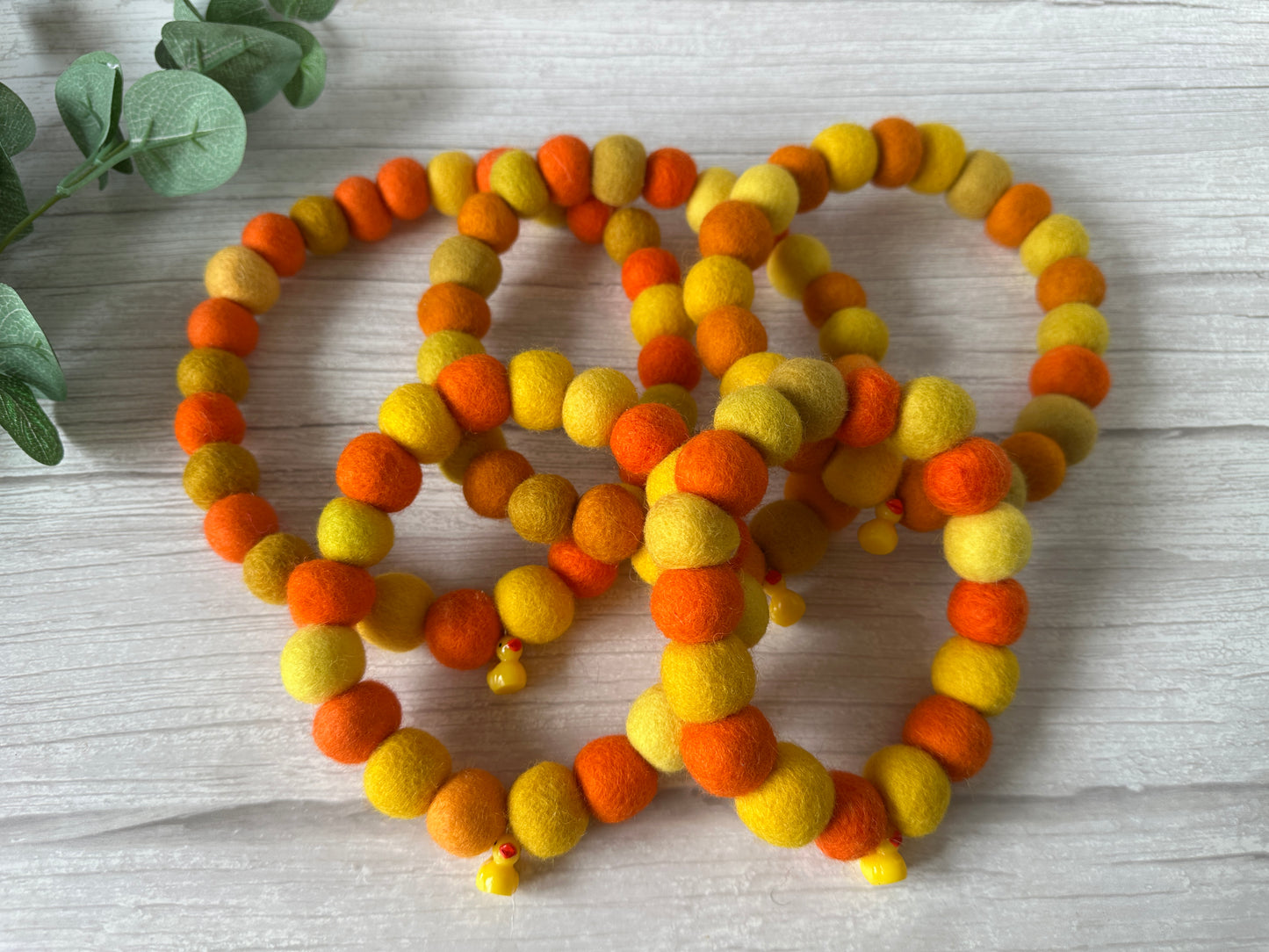 The PomPom Pet Collar - Rubber Ducks by Crafts by Kate features four beaded bracelets in orange and yellow felt, displayed on light wood—ideal as an elasticated pet collar. A sprig of green leaves graces the upper left corner.