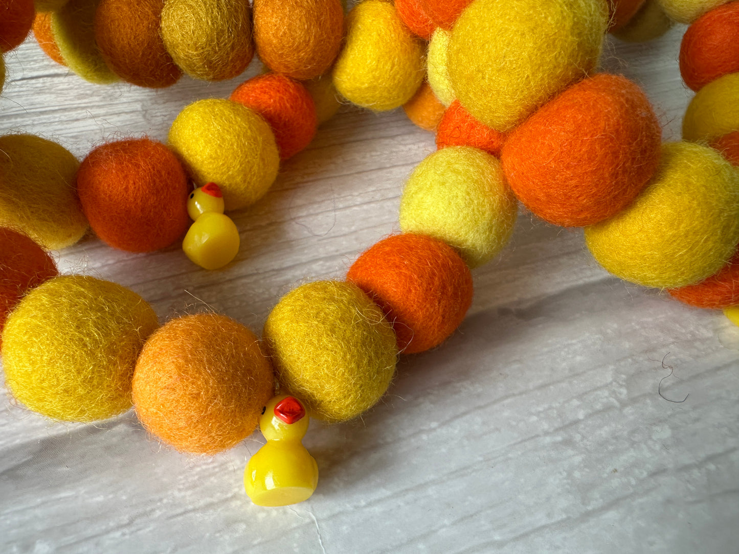 Close-up of the PomPom Pet Collar - Rubber Ducks by Crafts by Kate, featuring yellow and orange felt balls and small plastic duck charms, displayed on a light wooden surface.