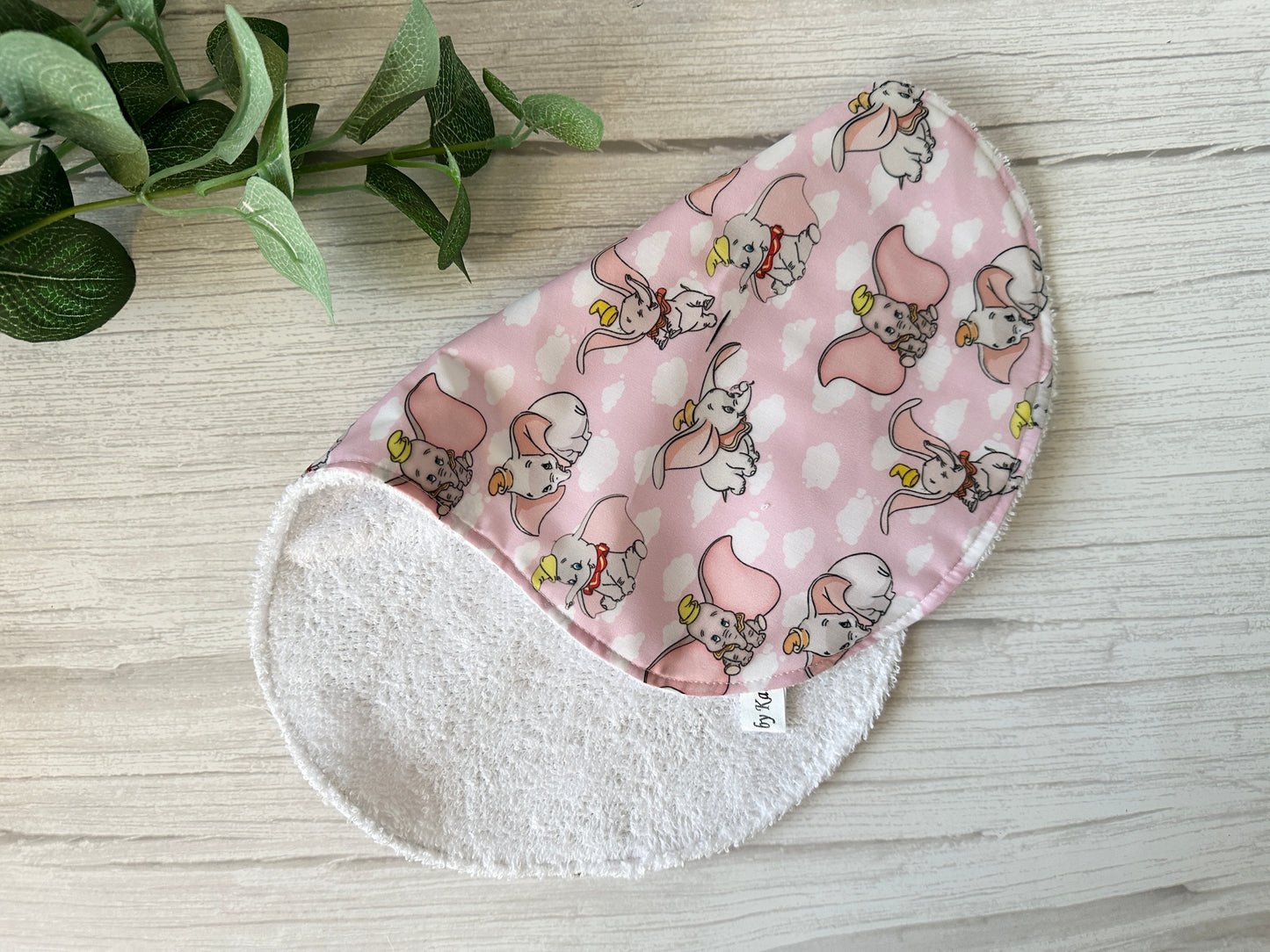 The Cotton & Terry Towelling Baby Gift Set - Dumbo in pink by Crafts by Kate is displayed on a light wood surface. Made from handcrafted ultra-soft cotton with a plush white terry underside, the cloth showcases cartoon elephants and clouds. A small plant nearby completes this charming set.