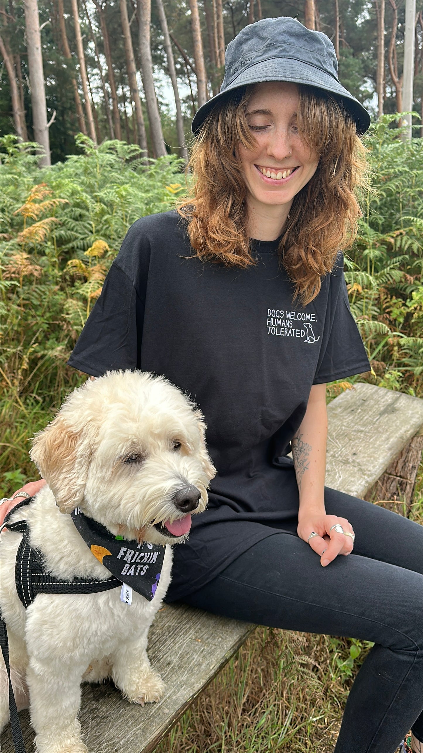 A smiling person with wavy brown hair, wearing a black "Dogs welcome" t-shirt by Crafts by Kate and a bucket hat, sits on a bench outdoors beside a fluffy white dog. Green trees and foliage fill the background.