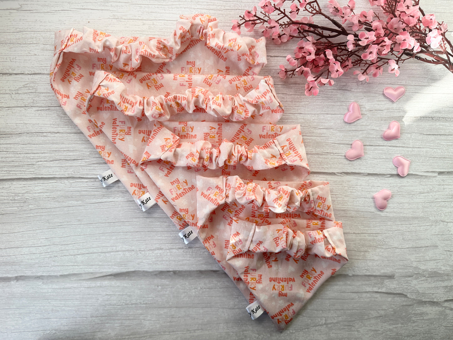Four "Cotton Pet Scrunchie Bandana - My Fluffy Valentine" by Crafts by Kate, with "my FURry valentine" text in red and orange, are displayed on a light wooden surface. Pink flowers and six pink heart-shaped objects add to the charming scene.