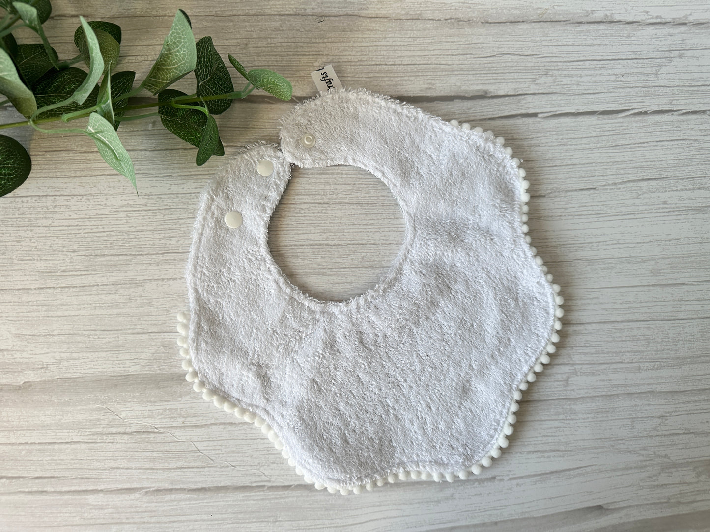 Crafts by Kate's Cotton & Terry Towelling Baby Gift Set - Floral Bambi, featuring a white textured bib with scalloped edges and pom-pom trim, lies on a light wooden surface. A green leafy plant in the top left corner completes this perfect baby gift set addition.