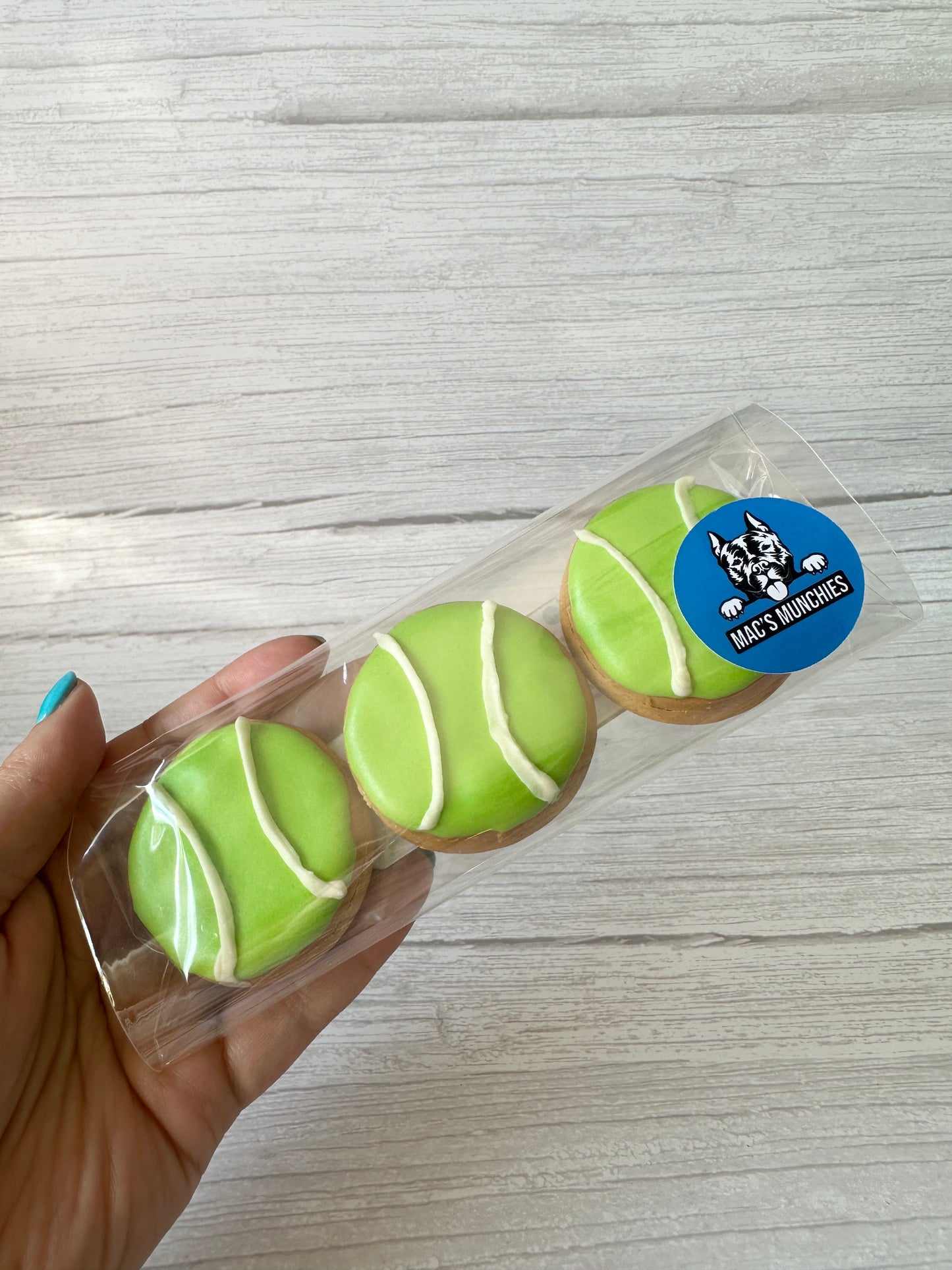 A hand holds a clear package with three round cookies decorated as green tennis balls with white icing. The blue sticker with dog logo and "MAC'S SNACKIES" shows these are "Ball is Life - Arnie & Mac’s Goodies" from Crafts by Kate.
