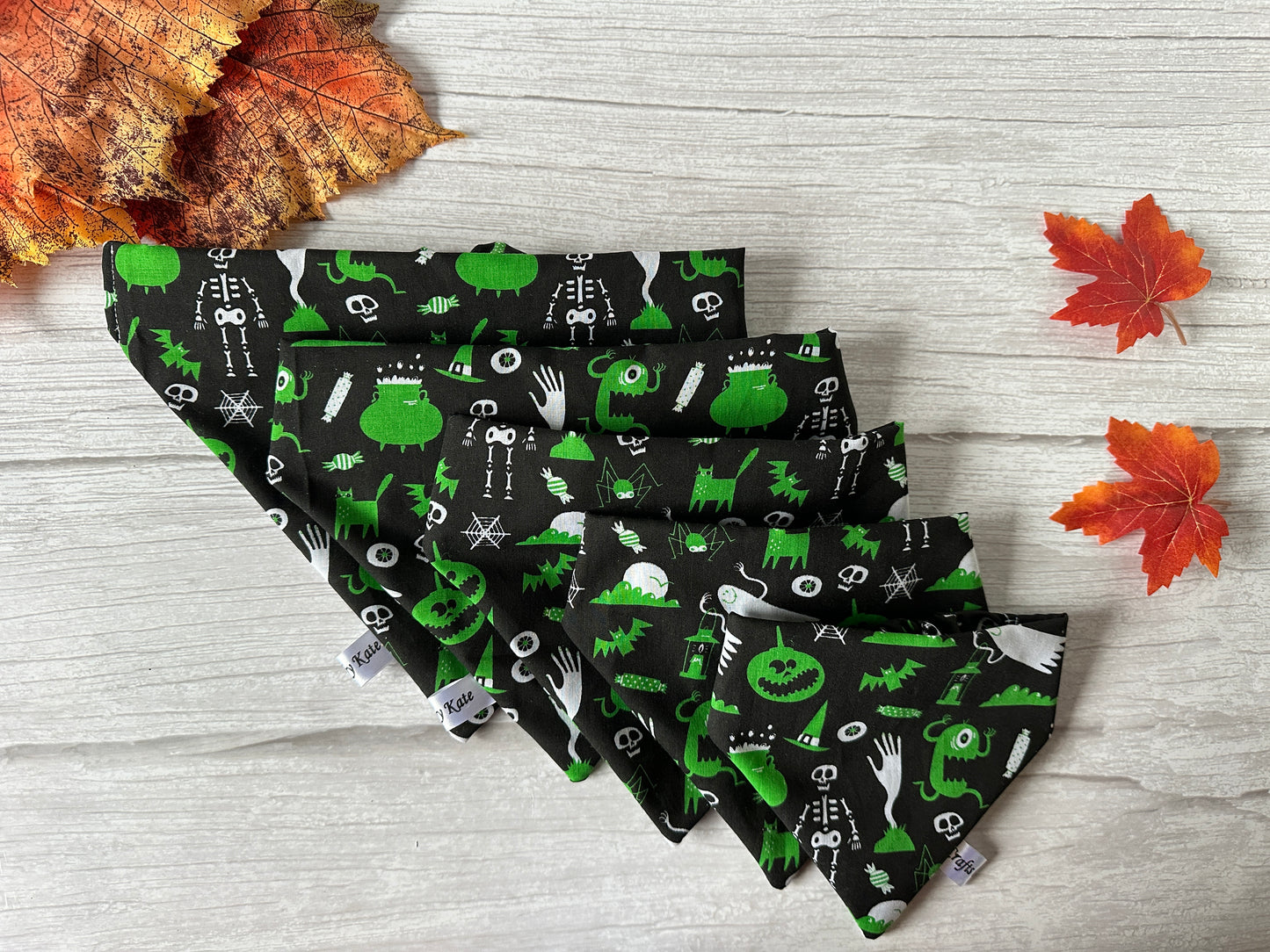 The Cotton Pet Scrunchie Bandana - Green Halloween by Crafts by Kate is handcrafted from cotton with skeletons, pumpkins, and bats on green fabric. Available in various sizes and displayed with autumn leaves on light wood.