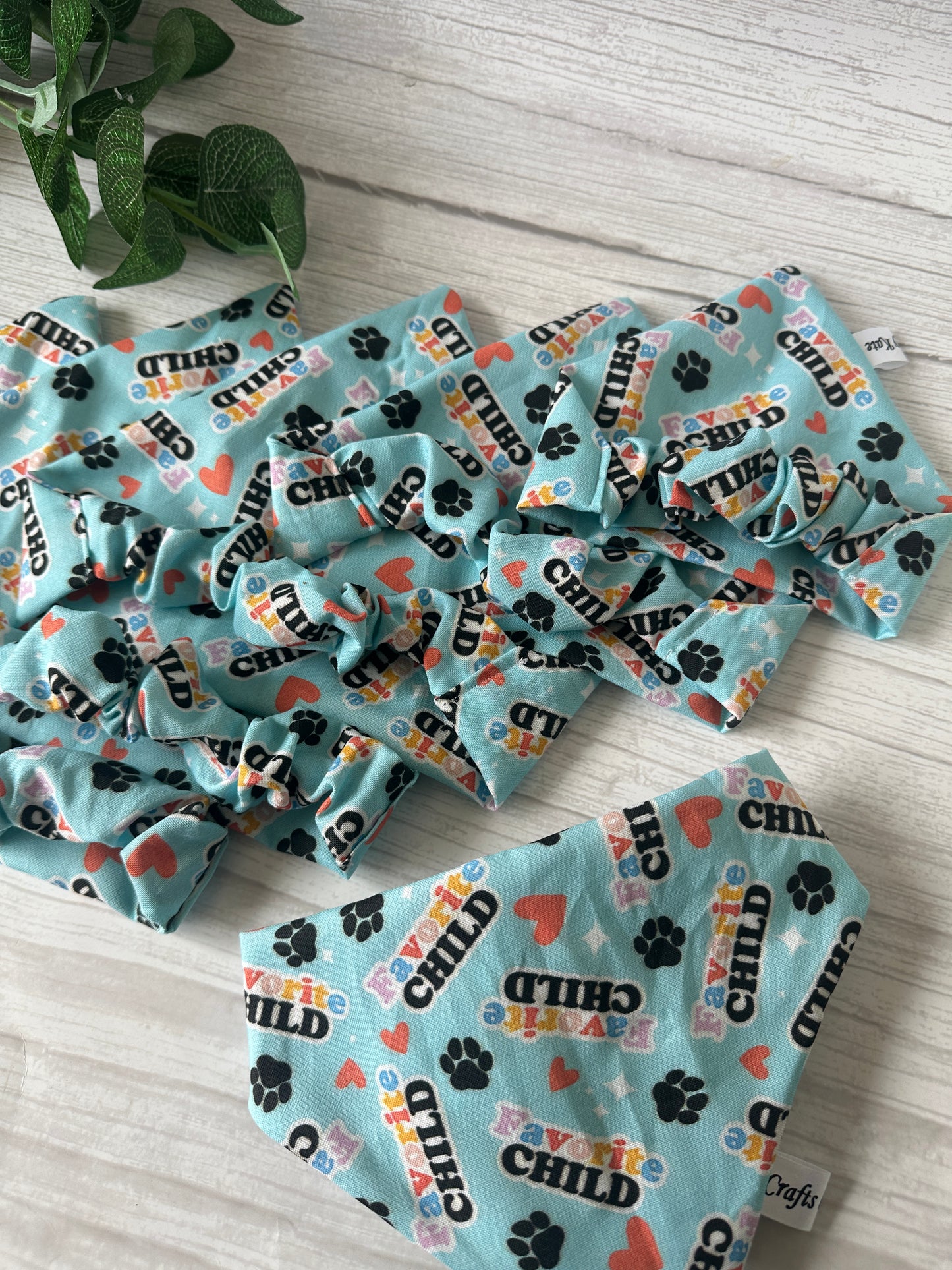 Crafts by Kate's Cotton Pet Scrunchie Bandana - Favourite Child features blue fabric adorned with "Favorite Child," hearts, and black paw prints. A green leafy plant decorates the upper left on a light wood surface.