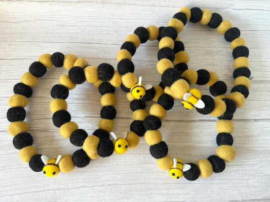 The PomPom Pet Collar - Bumble Bee by Crafts by Kate features black and yellow felt balls with a cute bee charm, displayed on a light wooden surface.