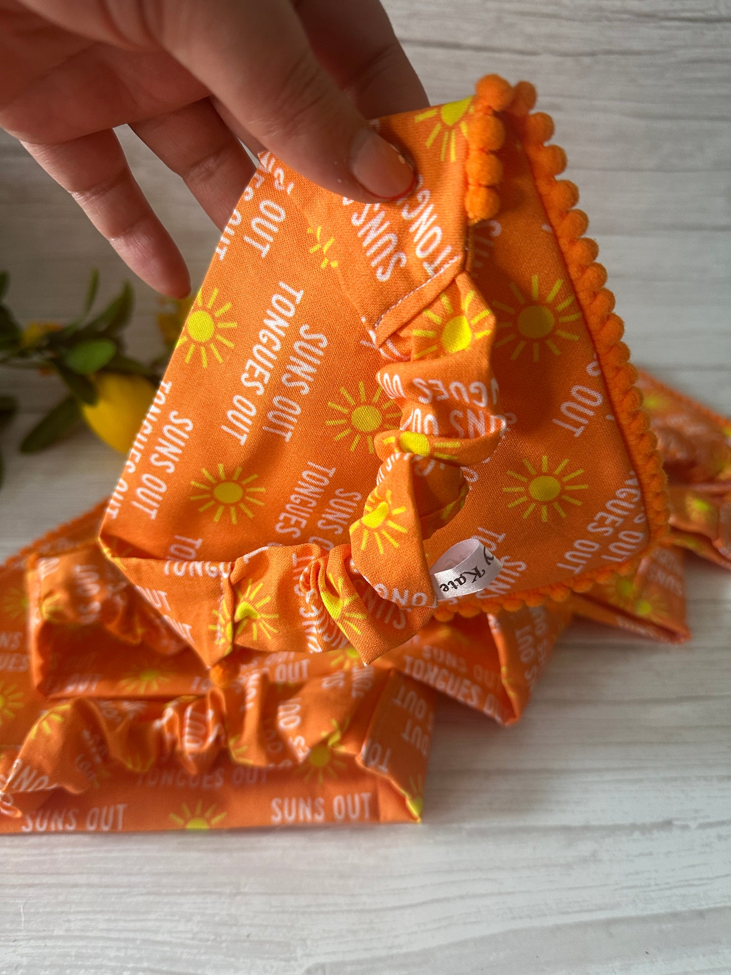 A hand holds the Crafts by Kate Cotton Pet Scrunchie Bandana - Tongues out, Suns out, made of bright orange fabric with yellow suns, "SUNS OUT TONGUES OUT" text, and an orange pom-pom trim. A leafy plant is in the background.