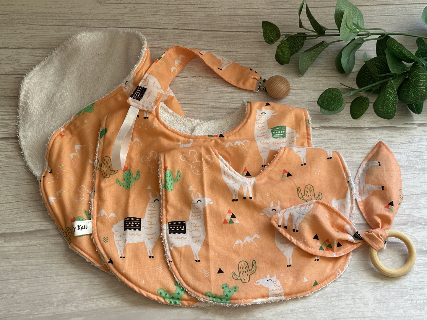 Crafts by Kate's Cotton & Terry Towelling Baby Set, "Llama Loves," features peach-colored, ultra-soft bibs with llama and cactus designs and a bamboo teething ring with a fabric knot. Placed on a wooden surface beside green leaves, this handcrafted set blends charm and practicality.
