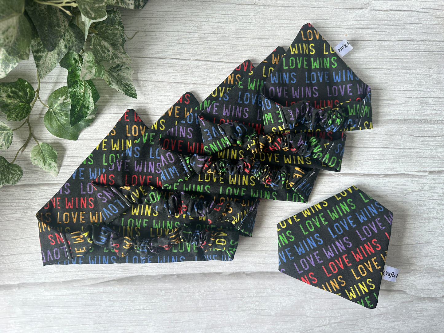 The Cotton Pet Scrunchie Bandana - Love Wins by Crafts by Kate features rainbow "LOVE WINS" on black cotton, celebrating the LGBTIQA+ community. Handcrafted accessories are styled on a light wooden surface with green leaves in the corner.