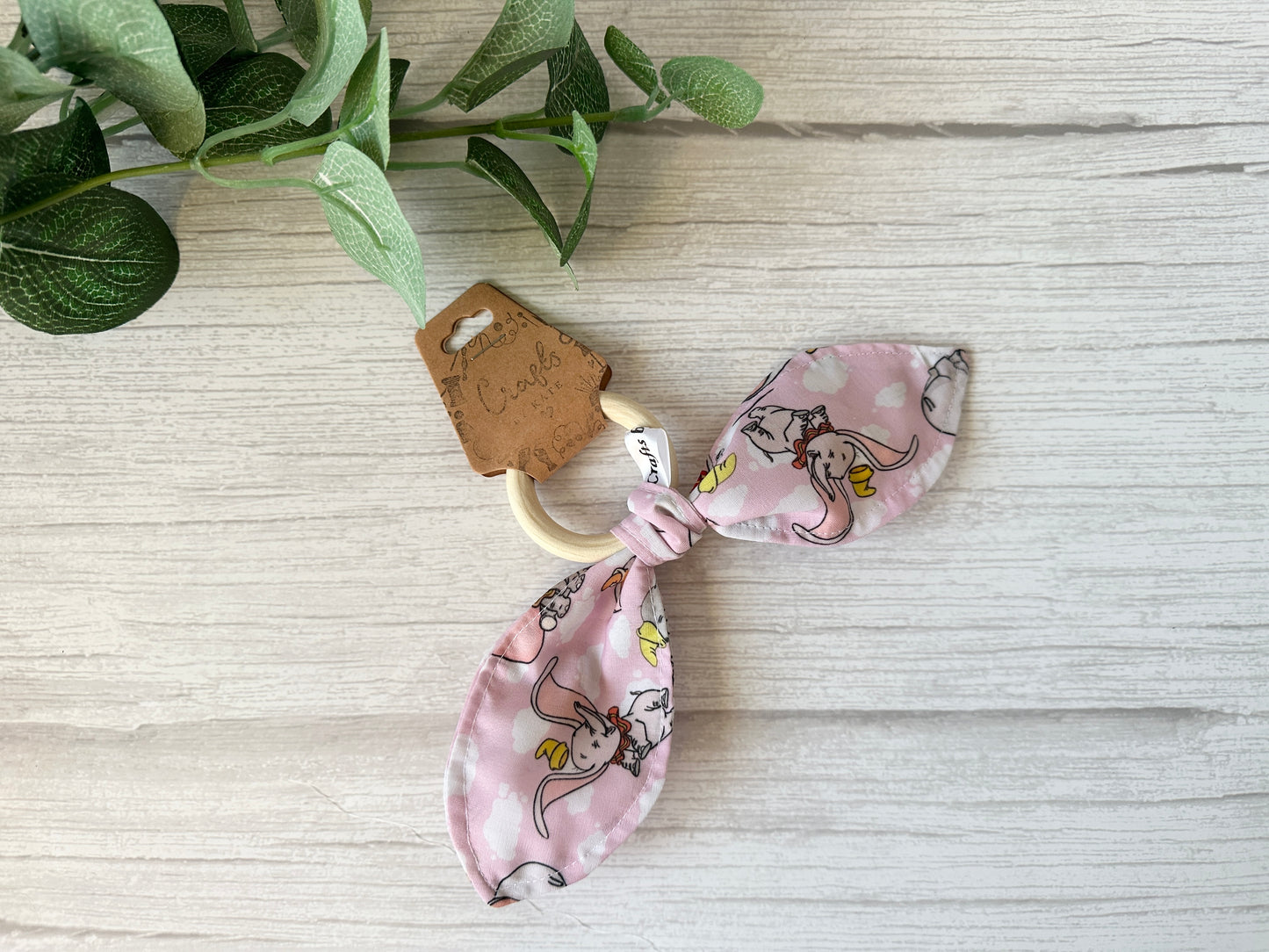 The charming Cotton & Terry Towelling Baby Gift Set - Dumbo in pink by Crafts by Kate features a pink fabric teether with a bamboo ring on a white wooden surface. Ultra-soft cotton shows playful cartoon cows and ducks, highlighted by a small green leafy branch for a natural touch.