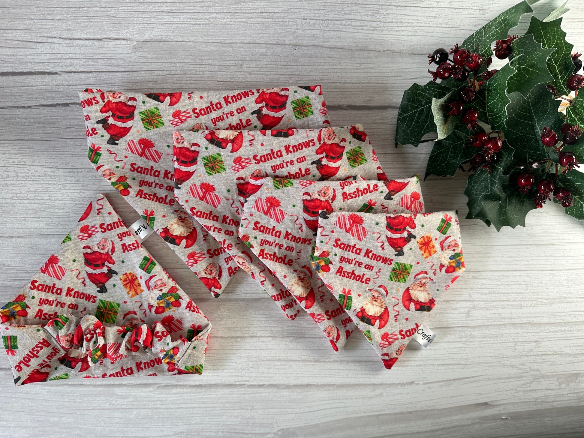 The Cotton Pet Scrunchie Bandana - Santa Knows by Crafts by Kate features Santa illustrations and playful text on festive fabric, styled on a light wooden surface with holly and red berries.