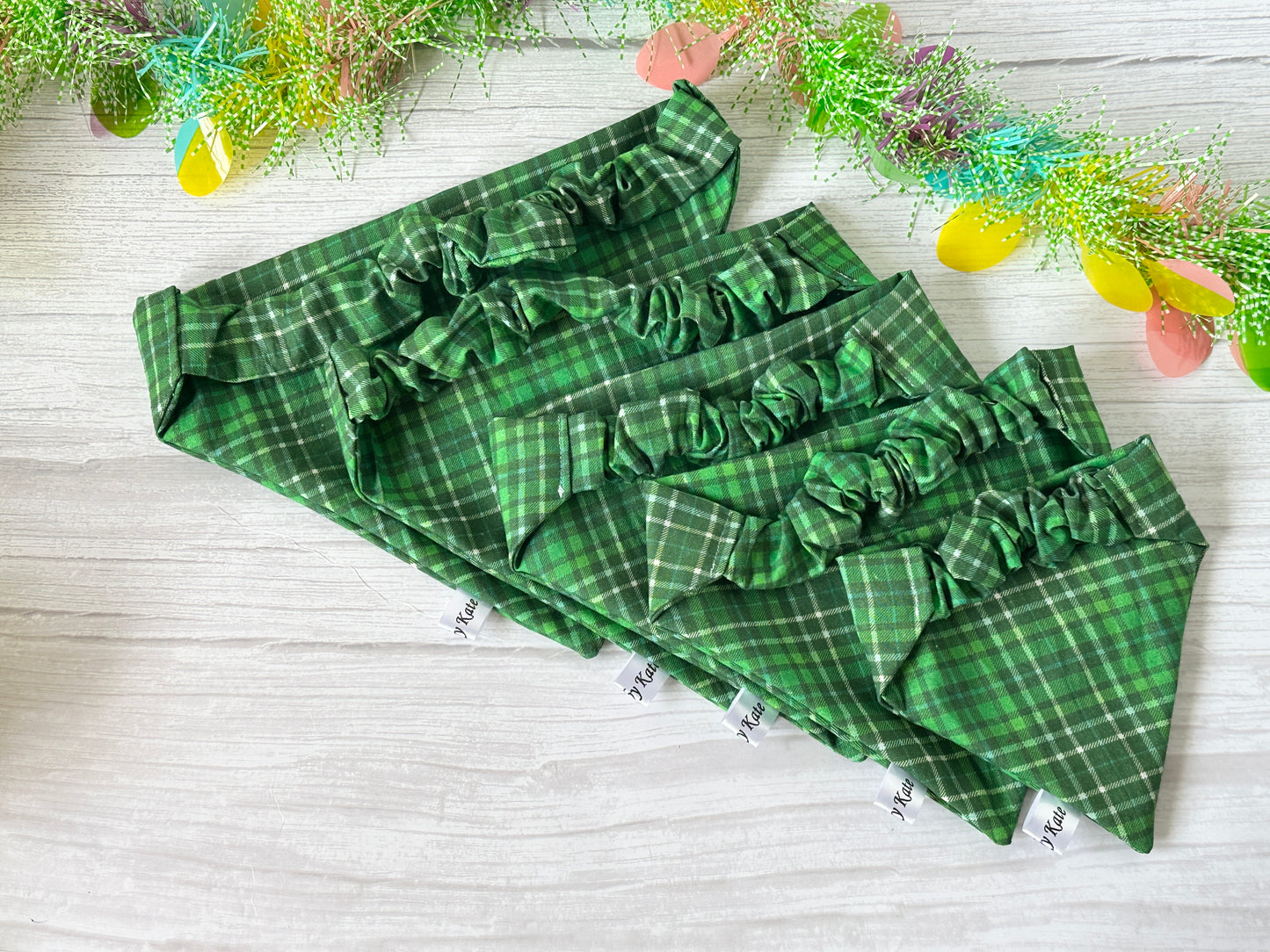 Five Crafts by Kate "Cotton Pet Scrunchie Bandana - Bluey & Bingo" in green plaid, made from spring tartan fabric with ruffled edges, are displayed on a light wood surface in a row from smallest to largest, with colorful foliage decorating the top edge of the image.