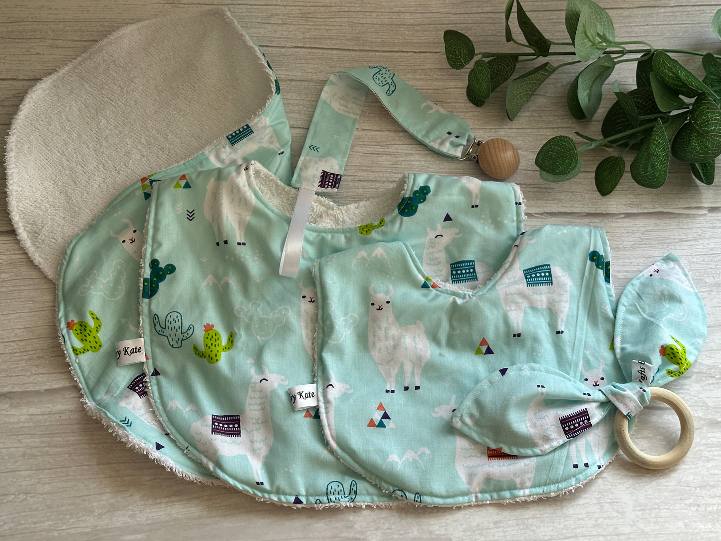 The charming "Llama Loves" baby set by Crafts by Kate includes handcrafted cotton bibs, a burp cloth, pacifier clip with llama and cacti patterns on light blue fabric, plus a wooden bamboo ring teether with a fabric knot, elegantly displayed on light wood.