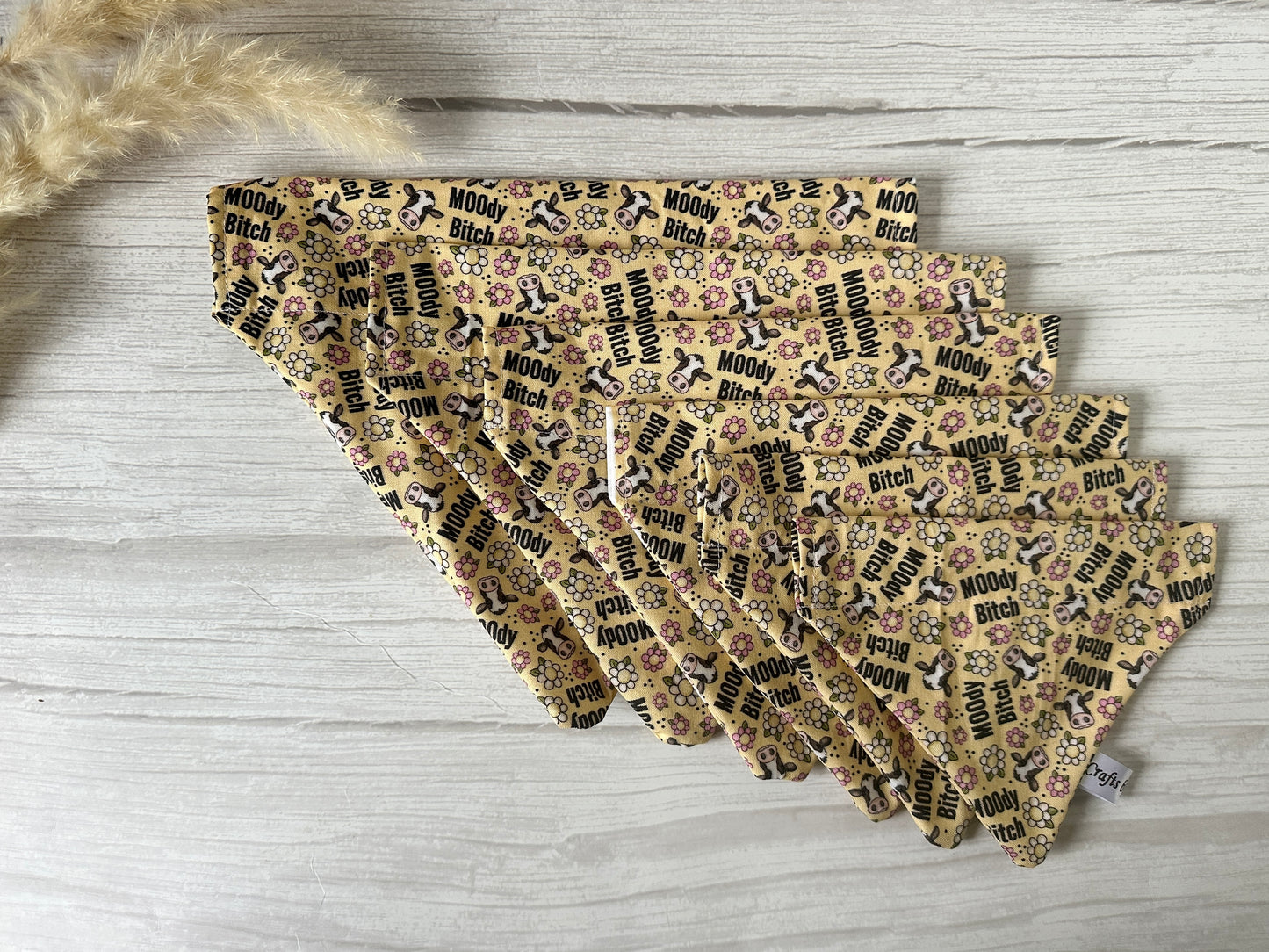 A spread of "Moody Cow" cotton pet bandanas by Crafts by Kate features handcrafted beige fabric squares with black text, playful pink flower doodles, and various animals on a light wood surface. Pampas grass is visible in the corner, enhancing its chic design.