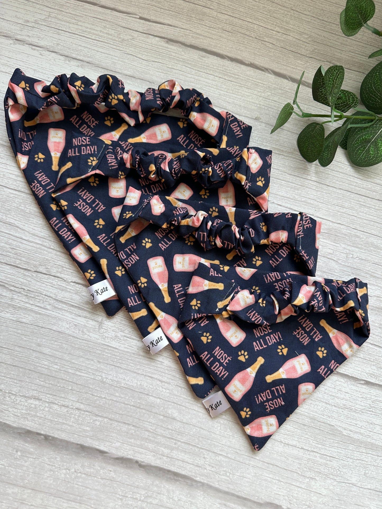 Four Cotton Pet Scrunchie Bandanas from Crafts by Kate, featuring paw prints and cartoon dog noses with "Nosé all day" text, are displayed on a wooden surface. A small plant is visible in the corner, enhancing this Gotcha Day display.