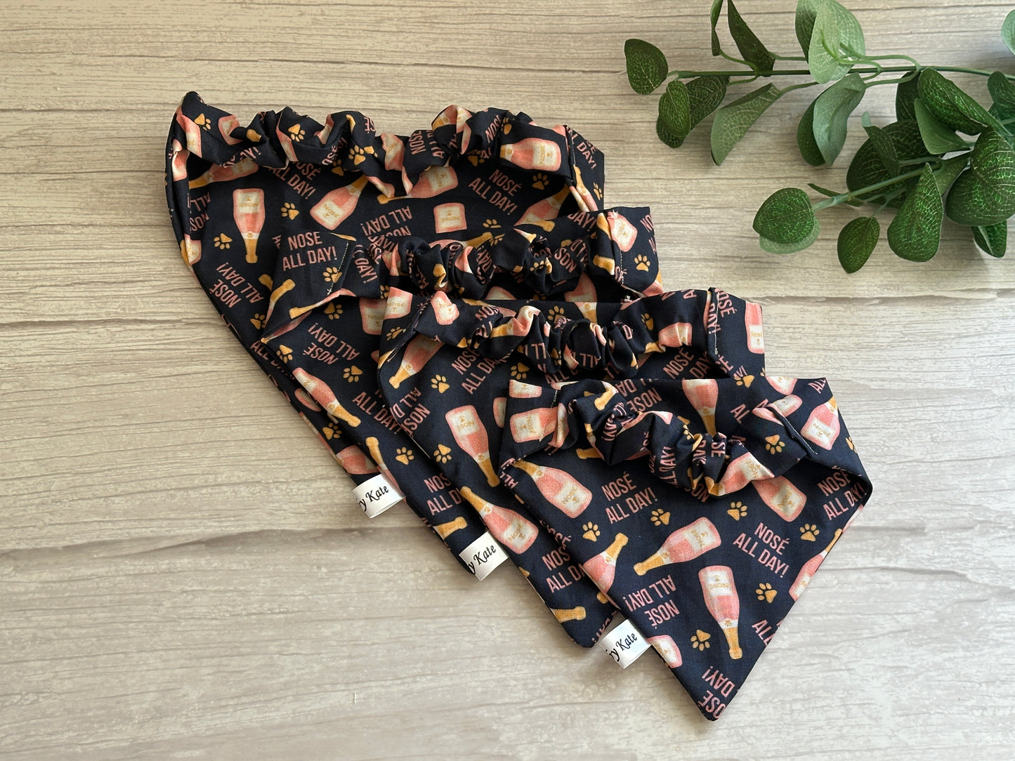 Three dark pet bandanas from Crafts by Kate, featuring "Nosé all day" with champagne glasses and paw prints, are elegantly placed on a wooden surface with green leaves nearby, ideal for your furry friend's Gotcha Day celebration.