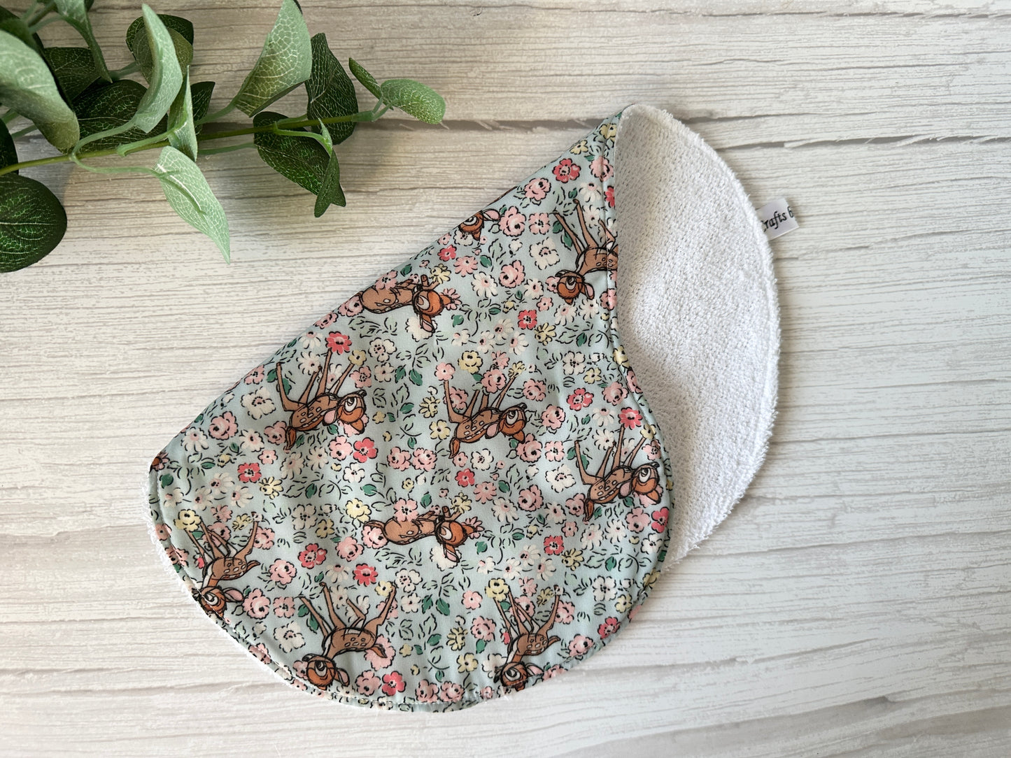 The Cotton & Terry Towelling Baby Gift Set - Floral Bambi by Crafts by Kate features an ultra-soft, floral-patterned oval swatch with animal illustrations on cotton and white terry cloth backing. It rests on a light wood surface with a green potted plant peeking from the top left corner.