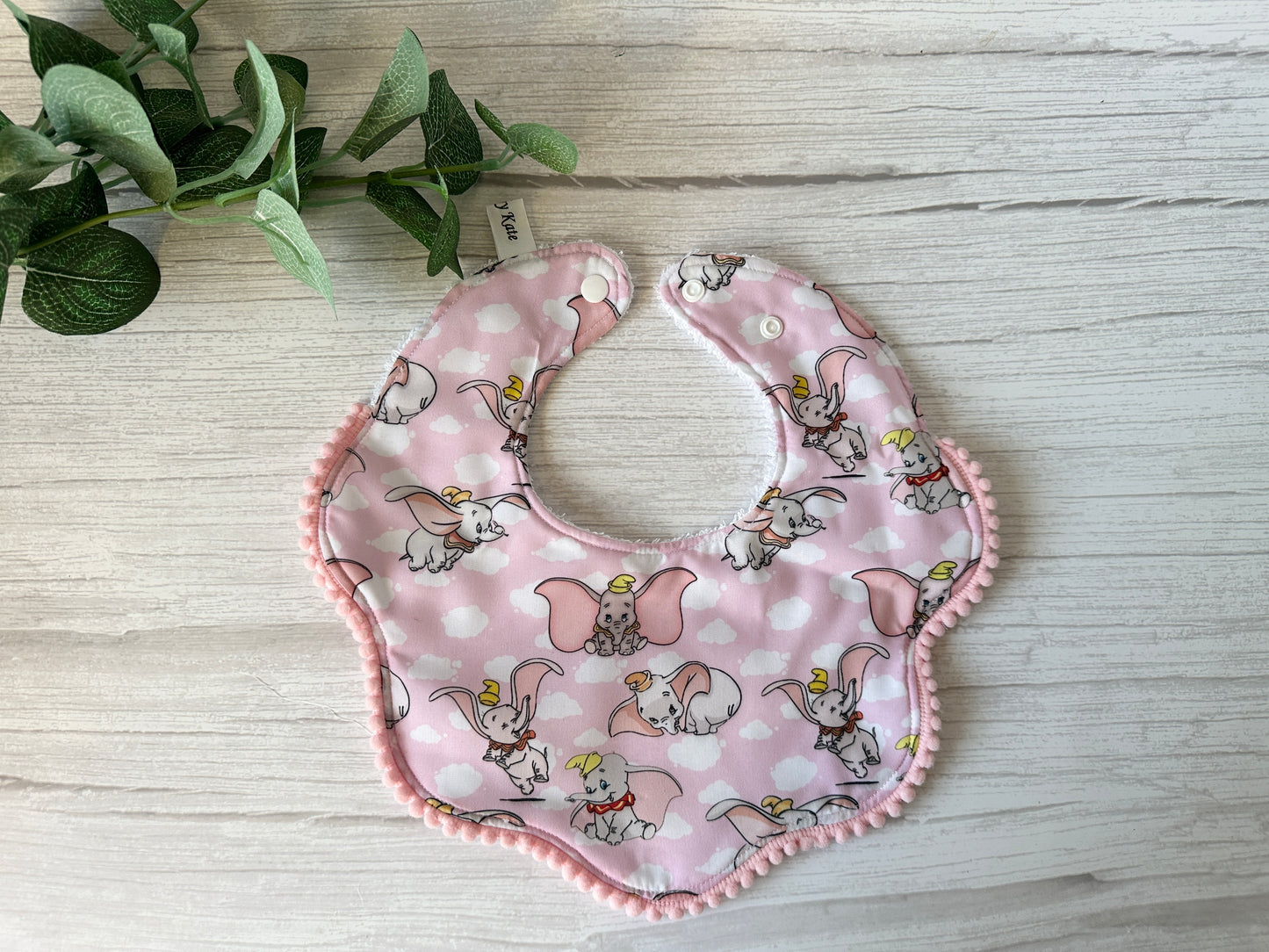 A pink baby bib from the "Cotton & Terry Towelling Baby Gift Set - Dumbo in pink" collection by Crafts by Kate features cartoon elephants in hats and wings, crafted from ultra-soft cotton with green-edged floral shape and a snap button, displayed on a light wood surface.