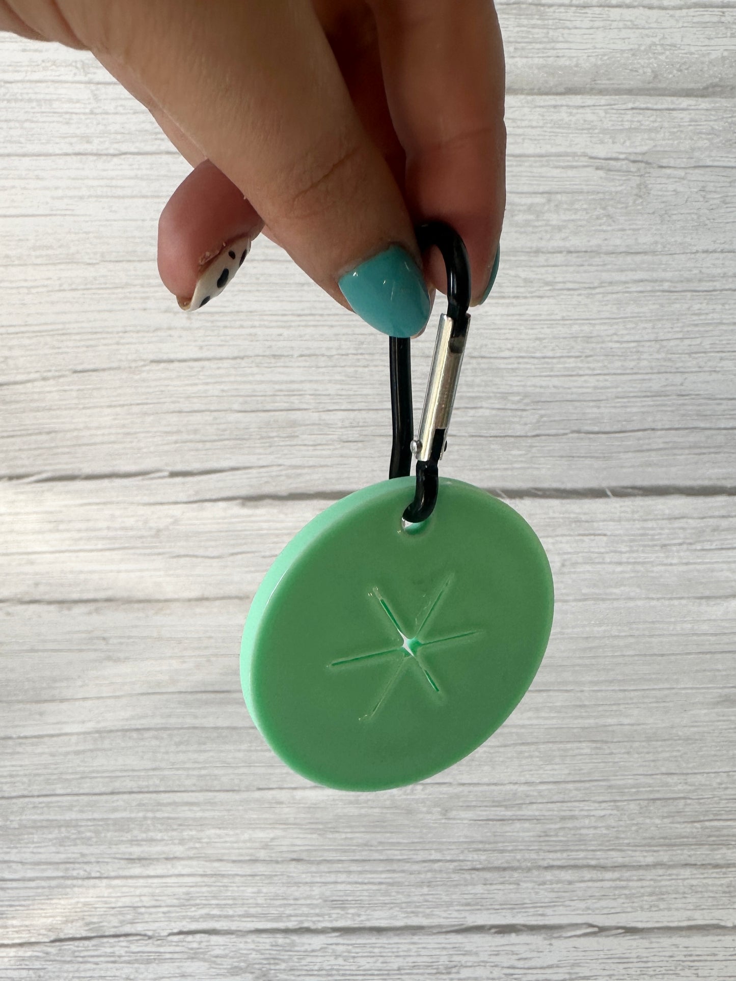 A hand with teal-painted nails holds Crafts by Kate's Ball is Life - Arnie & Mac’s Goodies, a green circular treat holder with an asterisk-shaped indentation and black carabiner—great for dog treats—against light wood.