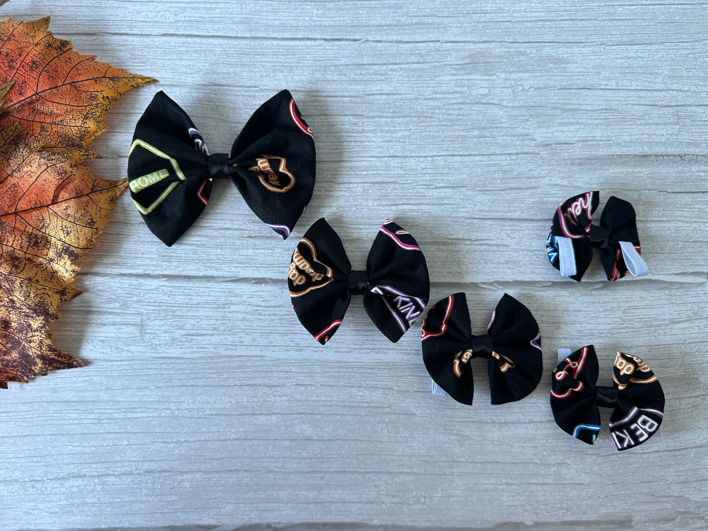 Five "Glow in the dark Signs" cotton pet bows by Crafts by Kate, each with a collar attachment, are arranged in a row on light wood beside an autumn leaf.