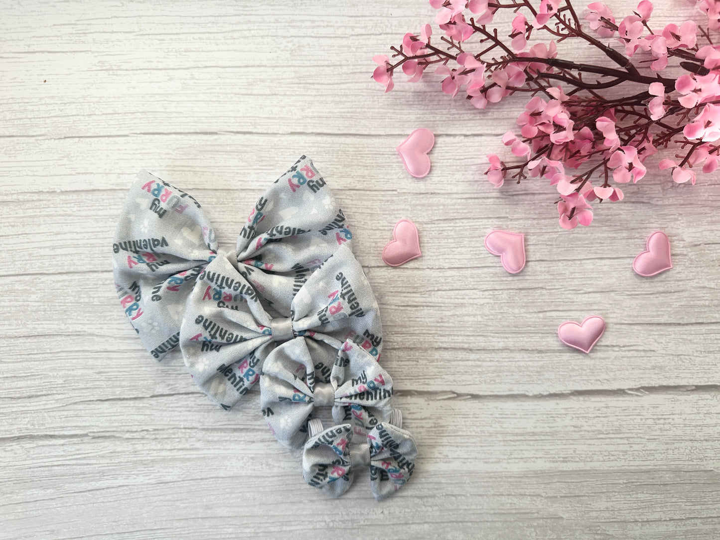 Gray, soft cotton hair bows with colorful text rest on light wood. Nearby, Crafts by Kate's Cotton Pet Bow with collar attachment - My Fluffy Valentine is surrounded by pink hearts and flowers for a cozy Valentine's vibe.