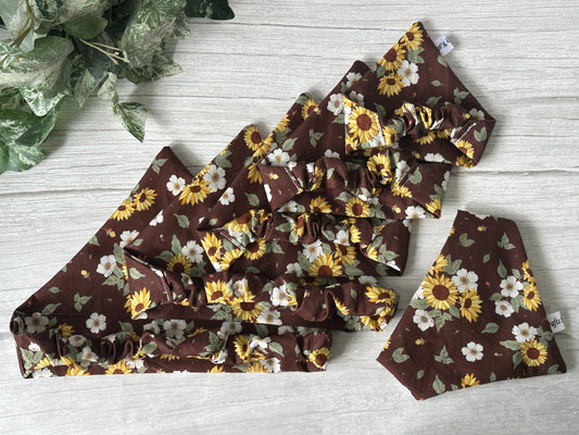 Five Cotton Pet Scrunchie Bandanas in the Sunflower Garden design by Crafts by Kate, featuring brown cotton with yellow and white sunflowers and daisies, are displayed on a light wooden surface next to green leafy plants.