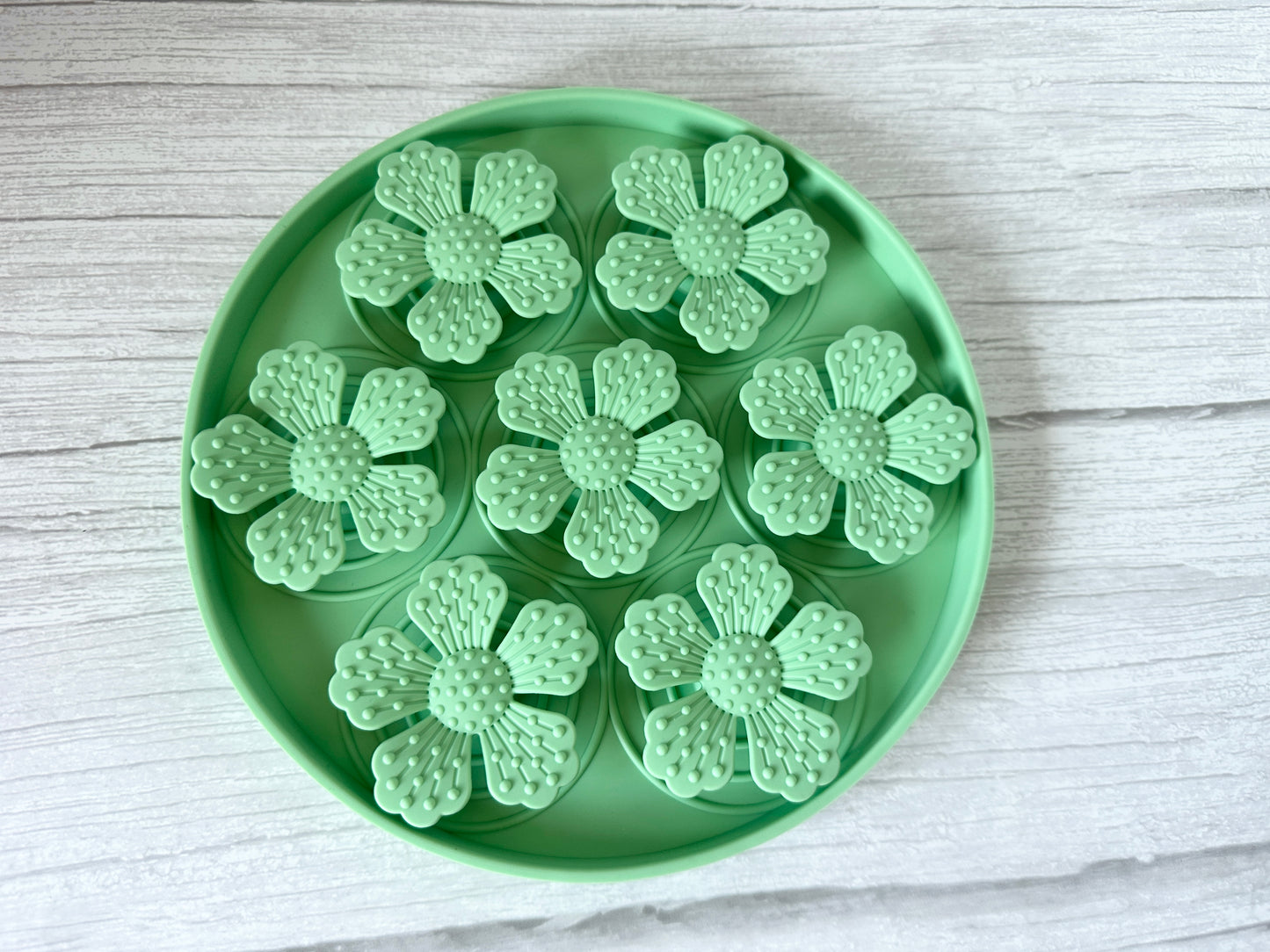 The round green Slow Feeder Pet Mat by Crafts by Kate features suction cups and holds seven mint-green, flower-shaped plastic items with detailed petals and textured centers. The tray sits securely on a light wood surface.