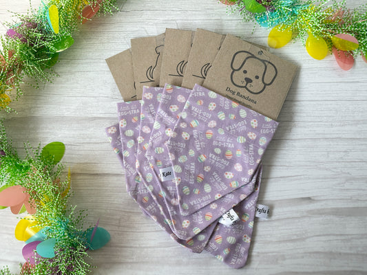 Several Crafts by Kate "Little bit Egg-stra" purple cotton dog bandanas with Easter patterns are arranged on a light wooden surface. Each bandana, featuring a collar attachment and a cardboard label with a dog illustration, is bordered by colorful, festive decorations.