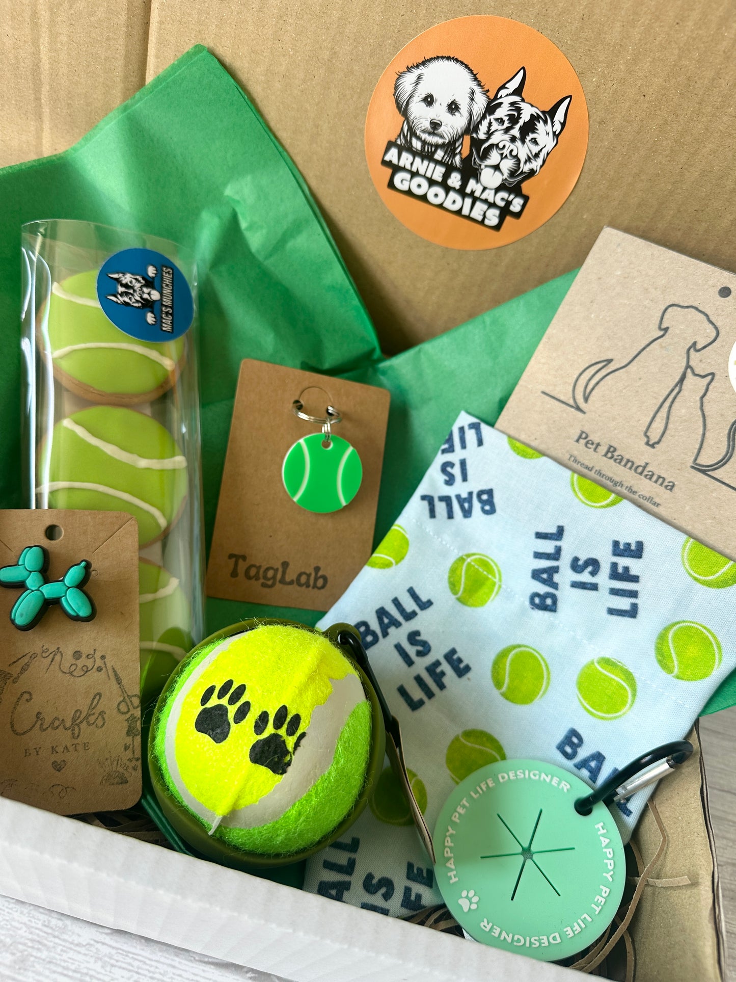 The Custom Order - Personalised Ball is Life Box by Crafts by Kate is a gift box with dog-themed tennis ball items: a chew toy, cookie, “L” or personalised bandana, tag, keychain, and stickers—all on green tissue paper.