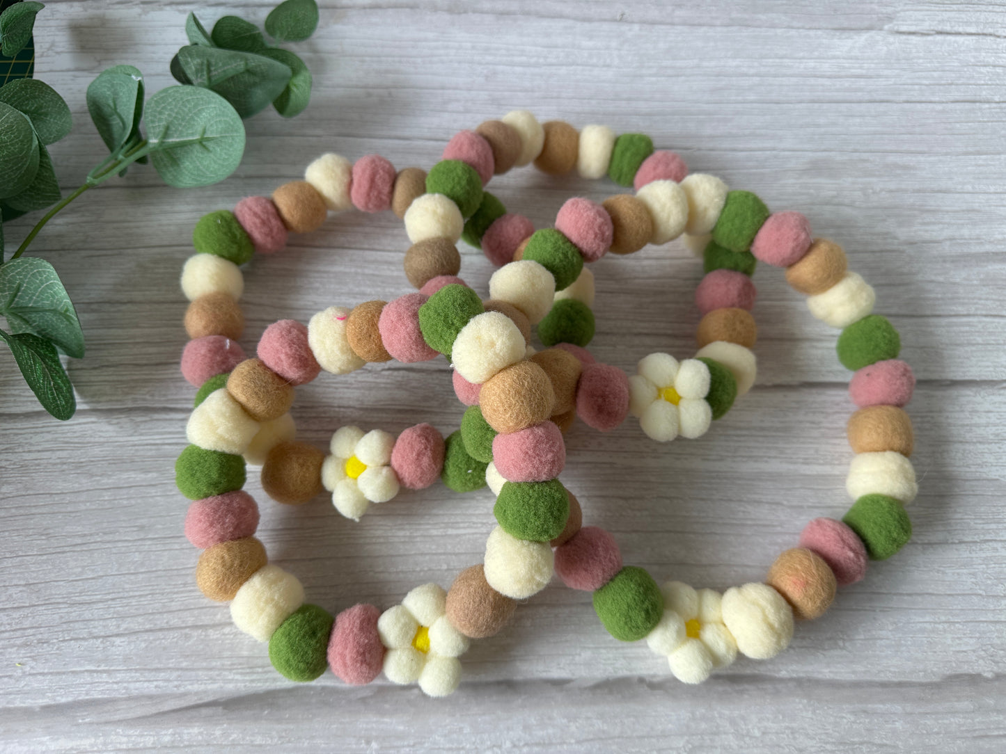 Three beaded felt ball bracelets in cream, green, pink, and brown with white felt flowers rest on light wood beside green leaves—perfect inspiration for the "PomPom Pet Collar - Fresh as a Daisy" by Crafts by Kate.