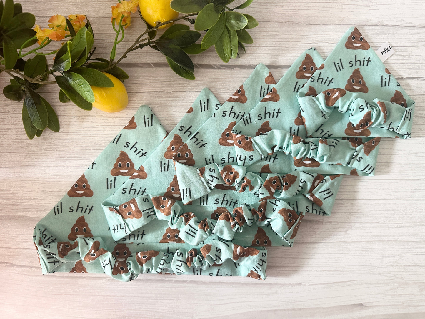 Four handcrafted Crafts by Kate Cotton Pet Scrunchie Bandana - Lil Shit sets, in aqua blue with a poop emoji and “lil shit,” are displayed with yellow flowers and lemons—fun, stylish headwear for your furry friend!.
