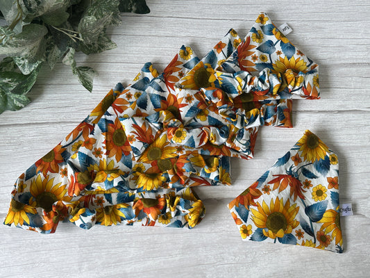 Six Cotton Pet Scrunchie Bandanas in the Sunflower Vibes design by Crafts by Kate are displayed on a light wooden surface, accented with green foliage peeking from the top left.
