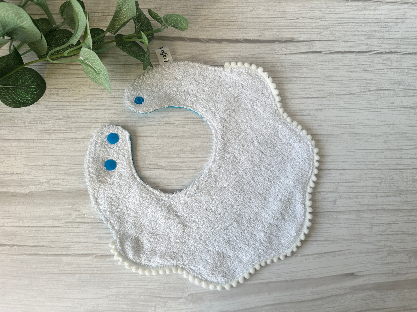A crafted "Gruffalo in blue" baby bib from Crafts by Kate's Cotton & Terry Towelling Gift Set, with scalloped edges, blue snap buttons, and pom-pom trim, lies on a light wood surface. A sprig of green leaves rests in the top left corner. All made from ultra-soft cotton.