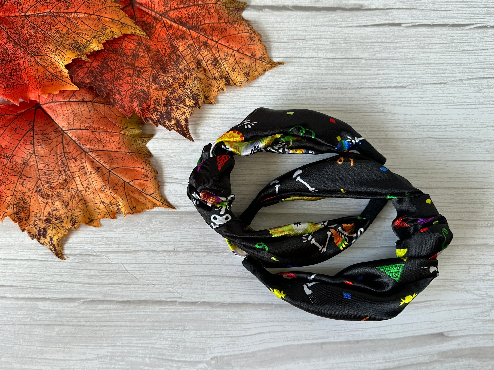 The Silk Knotted Headband - Day of the Dead by Crafts by Kate, featuring colorful skeletons and shapes, rests on a white wooden surface beside orange autumn leaves.