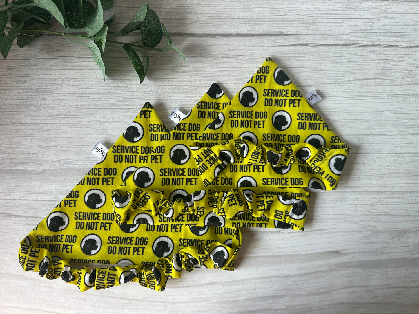 Three handcrafted Cotton Pet Scrunchie Bandanas - Anxious by Crafts by Kate, yellow with black "SERVICE DOG DO NOT PET" text and dog silhouettes, displayed on a white wooden surface. A green plant appears in the top left corner.