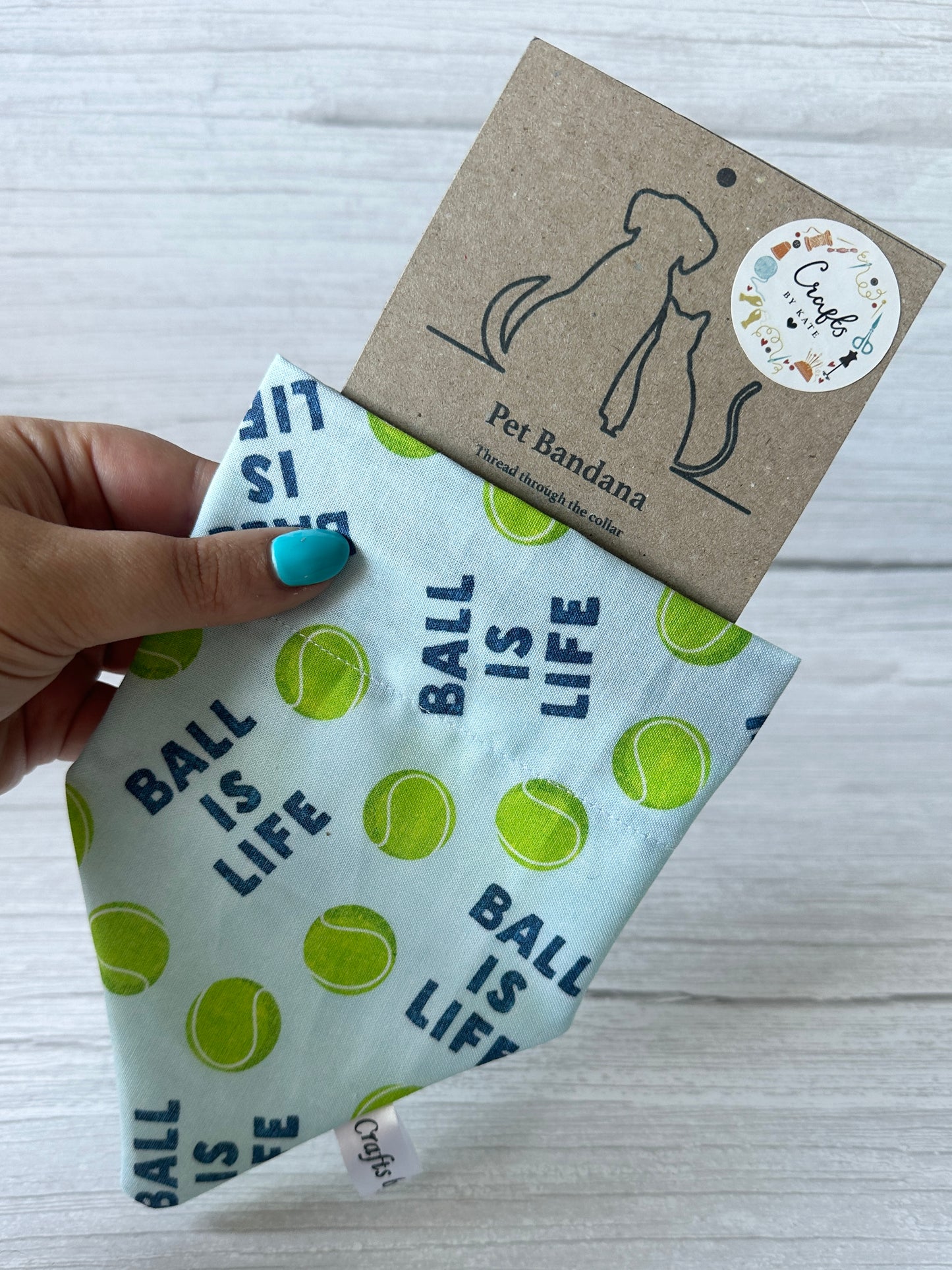 A hand holds a blue "Ball is Life - Arnie & Mac’s Goodies" pet bandana from Crafts by Kate, featuring green tennis balls and fun text, attached to "Pet Bandana" packaging with dog and cat outlines—ideal for your pup’s favorite treats.