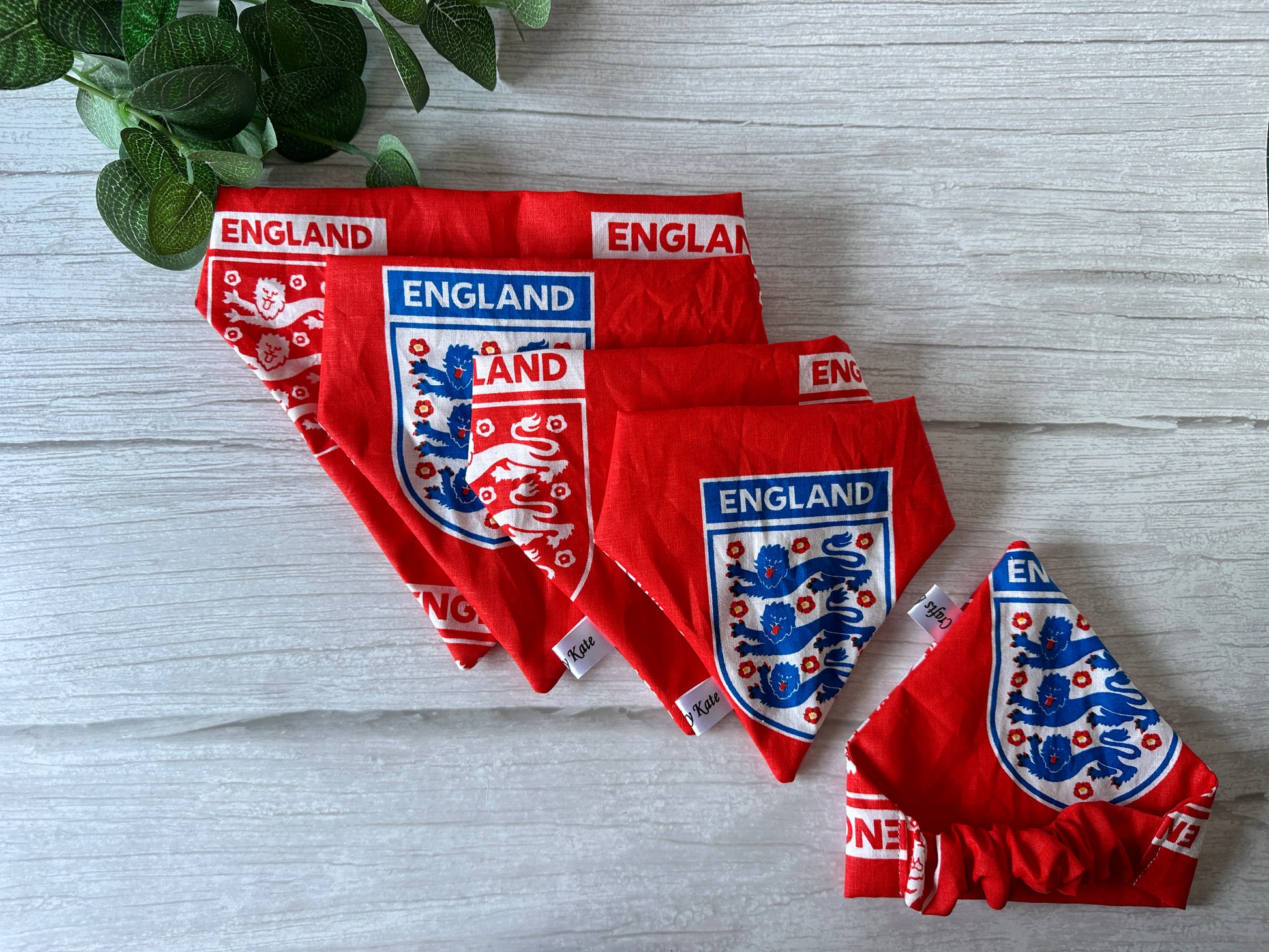 Four Crafts by Kate Cotton Pet Scrunchie Bandanas with the England Football Print are elegantly spread across a light wooden surface. A small green plant peeks in at the top left, adding a natural touch to this handcrafted display.