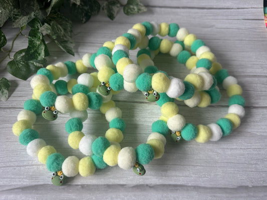 Three "PomPom Pet Collar - Frog Royalty" beaded bracelets by Crafts by Kate, featuring soft green, yellow, and white beads with frog face charms, are displayed on a white wooden surface with green leafy plants in the background.