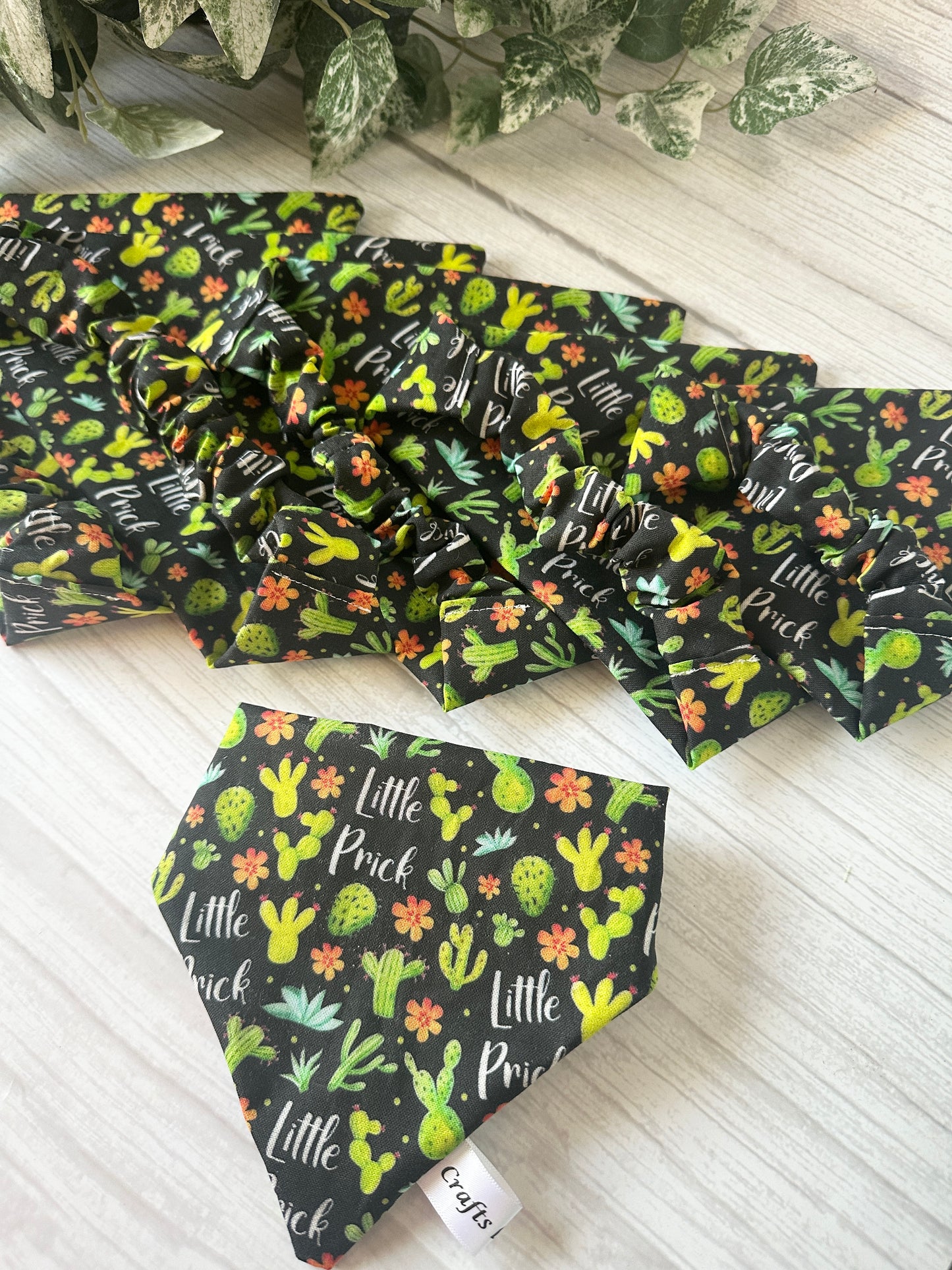 Several Crafts by Kate Cotton Pet Scrunchie Bandanas, featuring colorful cactus and flower designs with "Little Prick" text, are displayed on a light wood surface with greenery in the background.