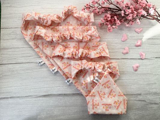 Three Cotton Pet Scrunchie Bandanas - My Fluffy Valentine by Crafts by Kate, made from fluffy Valentine fabric with pink tones and orange "my FUREVER valentine" text, are displayed on a light wooden surface with pink flowers and heart-shaped buttons, adding charm to the setting.